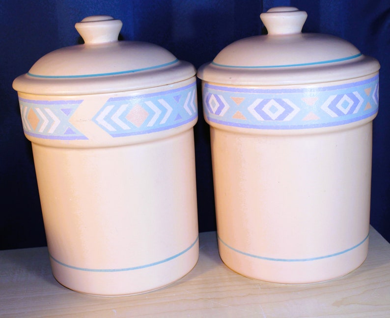 May include: Two ceramic canisters with lids, featuring a light pink base with a blue and white geometric pattern. The canisters are ideal for storing dry goods in the kitchen.