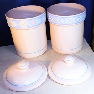 May include: Set of three ceramic canisters with lids. The canisters are white with a blue and pink geometric pattern around the top. The lids are white with a blue trim.