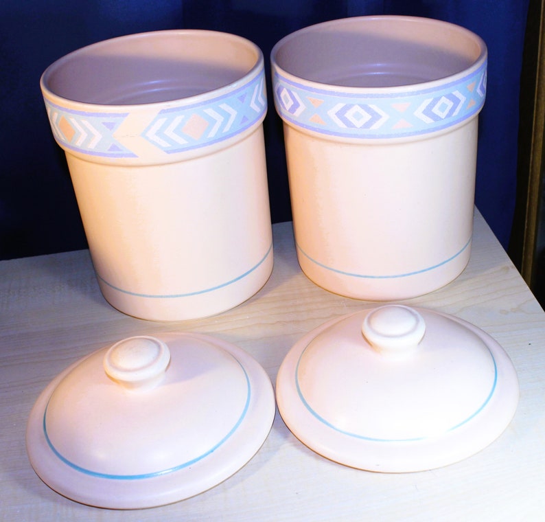 May include: Set of three ceramic canisters with lids. The canisters are white with a blue and pink geometric pattern around the top. The lids are white with a blue trim.