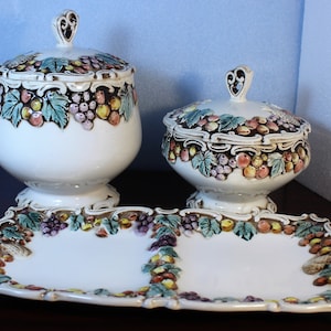 May include: A white ceramic serving set featuring a grapevine design. The set comprises two covered dishes and a rectangular serving tray. The dishes and tray are decorated with raised grapes, leaves, and vines in shades of green, brown, purple, and yellow.