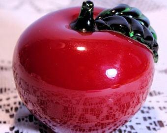 May include: A red glass apple with a green and black glass leaf on top. The apple is sitting on a white lace tablecloth.