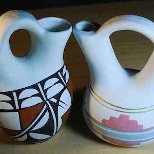 May include: Two white ceramic Native American pottery vessels with hand-painted designs. One vessel has a geometric pattern in black, white, and brown. The other vessel has a geometric pattern in gold, pink, and blue.