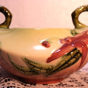 May include: A vintage ceramic bowl with two handles. The bowl is decorated with a floral design in shades of green, red, and white. The bowl is in good condition with no chips or cracks.