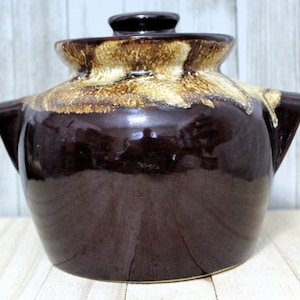 May include: A brown ceramic bean pot with a lid. The pot has a glossy finish with a lighter brown glaze around the rim and lid. It features two side handles and a small knob on the lid.
