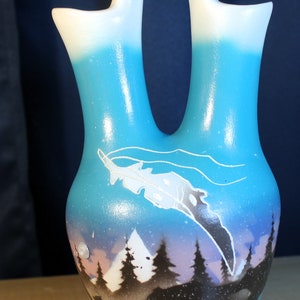 May include: A blue and white ceramic vase with a double spout and a painted design of a mountain scene with trees and a feather. The vase has a white rim and a light blue base.