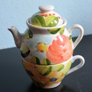 May include: A white ceramic teapot and teacup set with a floral design. The teapot has a lid and a spout. The teacup has a handle. The flowers are painted in shades of pink, yellow, blue, and green.