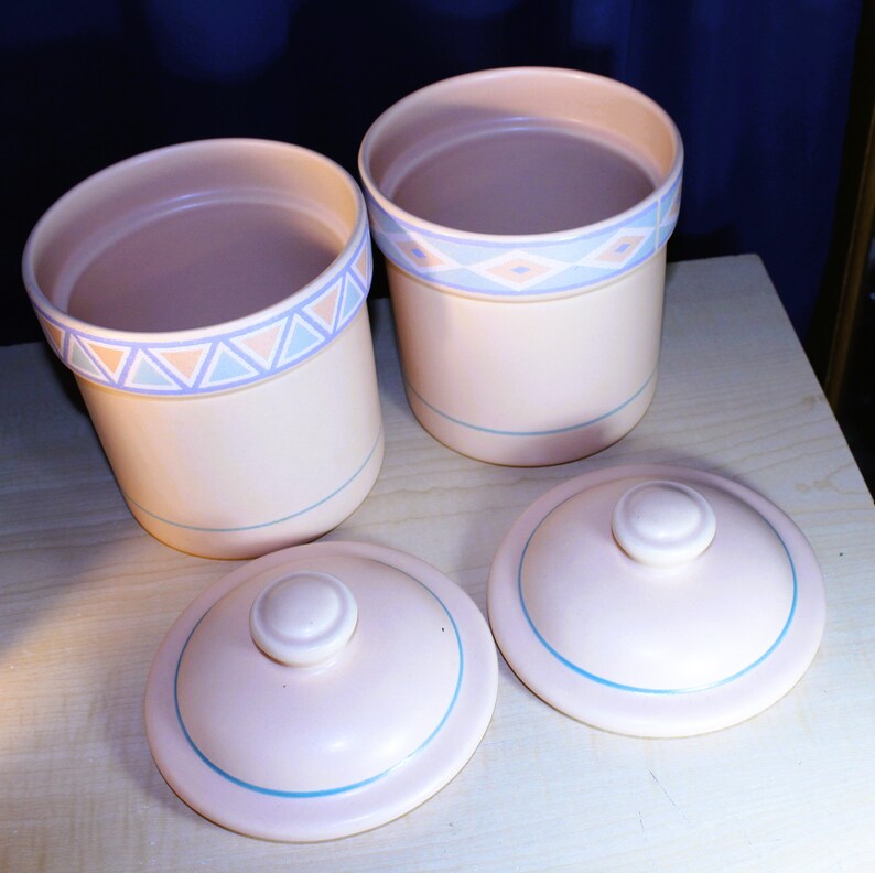 May include: Two ceramic canisters with lids, featuring a light pink base with a blue and pink geometric pattern around the top. The lids are also light pink with a blue trim.