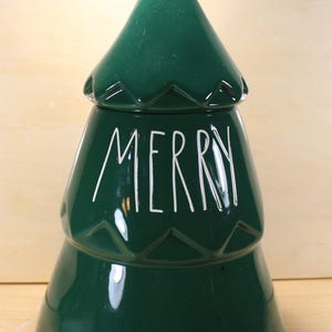 May include: A green ceramic Christmas tree-shaped container with a white star on top. The word "MERRY" is written in white on the front. The container has a glossy finish and is likely used for storage or decoration.