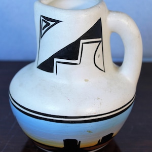 Op de afbeelding: Witte keramische kan met zwart-witte geometrische ontwerpen en een blauwe en gele zonsondergangscène met zwarte silhouetten van gebouwen.