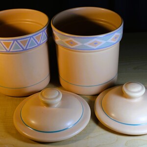 May include: Two ceramic canisters with lids, both with a light brown base and a blue and orange geometric pattern around the top. The lids are also light brown with a blue trim.