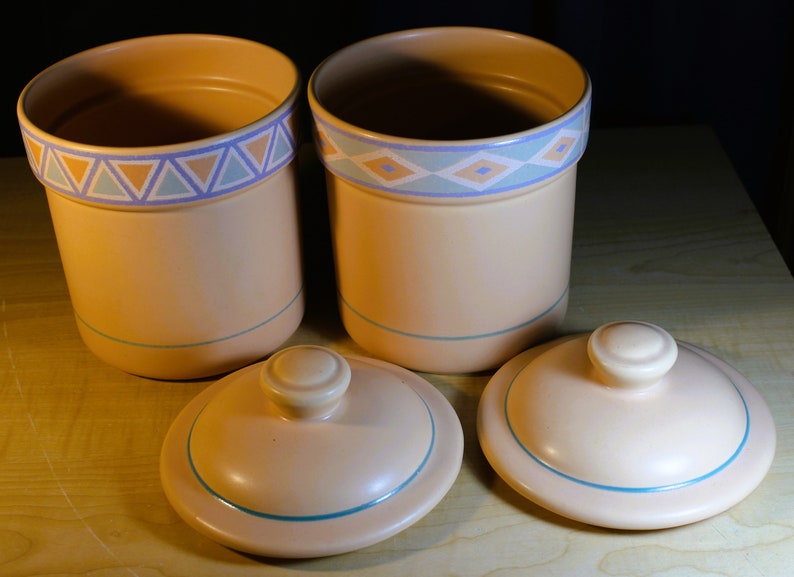 May include: Two ceramic canisters with lids, both with a light brown base and a blue and orange geometric pattern around the top. The lids are also light brown with a blue trim.