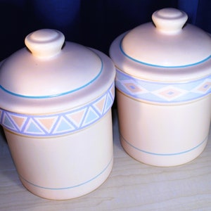 May include: Two ceramic canisters with lids, featuring a geometric pattern in shades of blue, pink, and white. The canisters are white with a light blue trim.