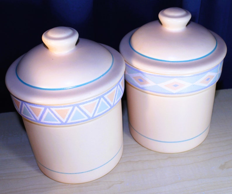 May include: Two ceramic canisters with lids, featuring a geometric pattern in shades of blue, pink, and white. The canisters are white with a light blue trim.