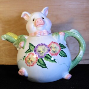 May include: A white ceramic teapot shaped like a pig with a pink bow. The teapot has a green handle and a corncob spout. The teapot is decorated with purple, pink, and yellow pansies.