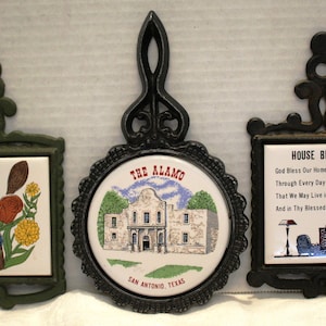May include: Three decorative trivets with cast iron frames. One features a floral design, another depicts The Alamo in San Antonio, Texas, and the third displays a "House Blessing" poem. Each trivet has a ceramic tile insert.