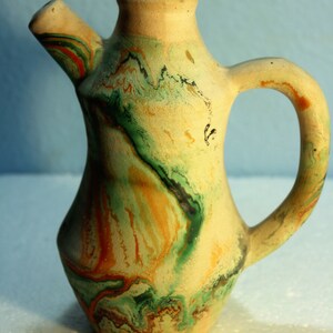 May include: A green, orange, and brown ceramic pitcher with a marbled design. The pitcher has a spout and a handle.