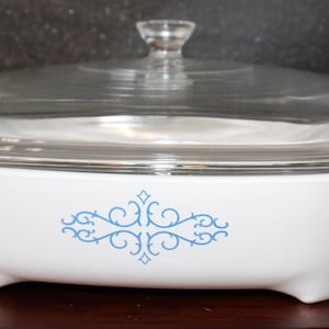 May include: White oval casserole dish with a glass lid and a blue floral design. The dish has a scalloped edge and three feet.
