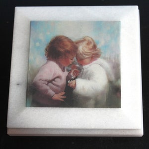 以下が含まれることがあります： 白い大理石の箱に、野原にいる2人の子供の絵が描かれています。左側の女の子はピンクのセーターを着て、右側の女の子は白いコートを着ています。2人はお互いを見つめ合い、小さな白い花束を持っています。