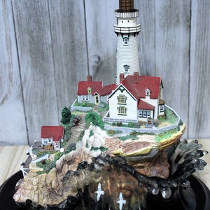 May include: A detailed miniature lighthouse scene with a white tower, red-roofed houses, and a rocky cliffside. The base features a stylized wave with three white seagulls. The lighthouse has a brown top and small windows.