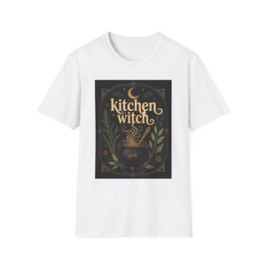 Op de afbeelding: Een wit T-shirt met korte mouwen en een centrale rechthoekige afbeelding op een donkere achtergrond. Het ontwerp toont de tekst 'kitchen witch' in een gestileerd lettertype boven een rokende ketel, omringd door botanische illustraties en een halve maan.
