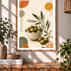 May include: A framed wall art print featuring a light-coloured bowl filled with green olives, accompanied by an olive branch with leaves. The composition includes abstract geometric shapes in orange, yellow, and green, set against a cream background.