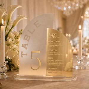 May include: A semi-circular acrylic table number sign labeled 'TABLE 5' in gold lettering, paired with a matching gold menu panel. The sign features a frosted finish and is mounted on a clear acrylic base, displaying a list of food items.