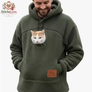 Op de afbeelding: Een olijfgroene fleece hoodie met een grote capuchon en een voorzak. In het midden van de borst zit een ronde opening waar de kop van een rood-witte kat doorheen steekt. Op de zak zit een kleine bruine rechthoekige patch met pootafdrukken.