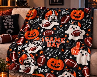 May include: A black fleece throw blanket featuring a repeating pattern of white ghosts wearing football helmets, orange jack-o'-lanterns, brown footballs, and the text 'GAME DAY' and 'Boo!' in orange and white lettering across the fabric.