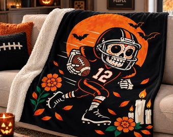May include: A black sherpa-lined throw blanket featuring a graphic of a skeleton football player wearing a helmet and jersey with the number 12. The design includes a large orange moon, flying bats, marigold flowers, and lit candles on a black background.