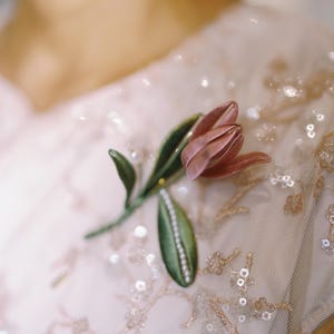 May include: A fabric tulip brooch featuring a dusty pink flower head and two green leaves. One leaf is accented with a row of small white beads. The brooch is pinned to a light pink sheer garment decorated with matching pink floral embroidery and sequins.