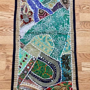 May include: A rectangular table runner featuring a patchwork design of various fabrics in shades of green, blue, red, and gold. The piece is embellished with intricate embroidery, sequins, beads, and small mirrors, finished with a dark border.