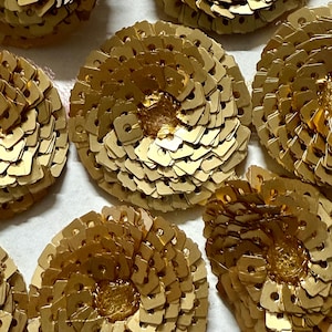 May include: Close-up of several golden, flower-shaped embellishments. Each flower is composed of layered, small, square-shaped pieces with a central, textured, golden core. The flowers are arranged on a light-colored surface.