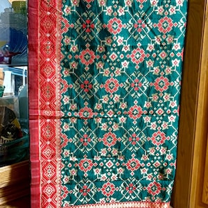 May include: A green and red patterned textile with a detailed geometric design. The fabric features a repeating pattern of red and white shapes on a dark green background. The edges are trimmed with a red border.