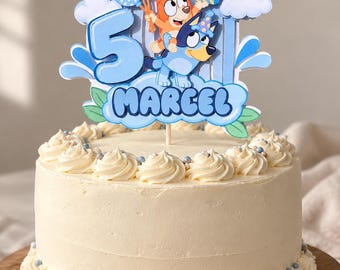May include: A round cake with white frosting and a decorative topper featuring two cartoon dogs, a large blue number 5, and the name Marcel. The topper includes blue balloons, a yellow sun, clouds, and green leaves, all set against a light blue arch.