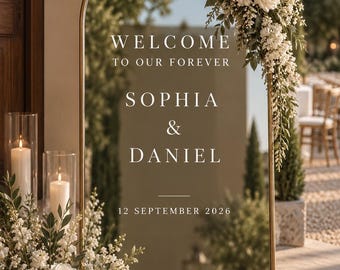 May include: An arched mirror wedding sign with a gold frame, featuring white text that reads WELCOME TO OUR FOREVER, SOPHIA & DANIEL, and 12 SEPTEMBER 2026. The mirror is decorated with white roses and greenery at the top right and bottom left corners.
