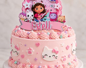 May include: A light pink frosted cake decorated with white cat faces, paw prints, and hearts. A large cake topper features the number 3, cartoon characters, and a pink banner with the name Estell written in white script lettering.