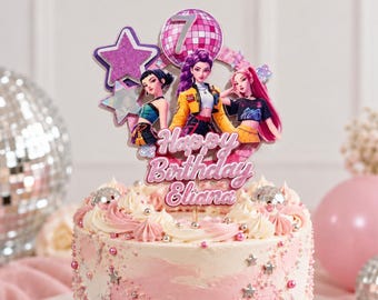 May include: A cake topper featuring three illustrated characters, a disco ball with the number 7, and purple stars. The text reads Happy Birthday Eliana in a pink script font. The topper is displayed on a pink and white frosted cake.