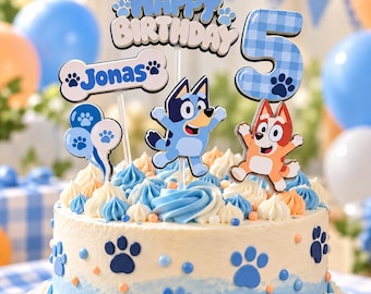 May include: A round birthday cake decorated with blue and white frosting swirls, paw print accents, and a cake topper set featuring the text Happy Birthday, the name Jonas on a bone shape, a large number 5, and two cartoon dog characters.