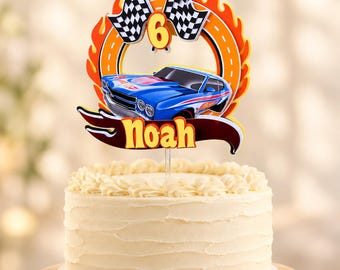 May include: A cake topper featuring a blue race car, the number 6, and two checkered flags set against an orange flame-shaped border. The name Noah is printed in yellow letters on a dark red banner at the base, all resting on a frosted cake.