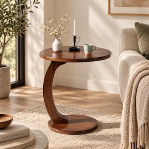 Puede incluir: Una mesa auxiliar redonda de madera marrón oscuro con una base en forma de C. La mesa presenta una superficie circular lisa sostenida por un único pedestal curvo que se conecta a una base plana y circular, proporcionando una estructura estable.