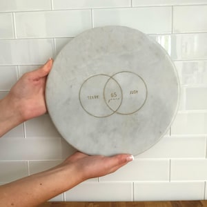 May include: A round white marble serving board featuring a gold-colored graphic of two overlapping circles. Inside the circles, the text reads TERRY, 65 years, and JUDY in a gold-colored font, centered on the smooth stone surface.