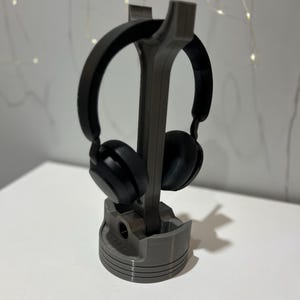 Könnte beinhalten: Ein Paar mattschwarze Over-Ear-Kopfhörer auf einem grauen, vertikalen Ständer in Form eines Motorkolbens. Der Ständer verfügt über eine zylindrische Basis mit horizontalen Rillen und einen zentralen, aufrechten Stützarm für die Kopfhörer.