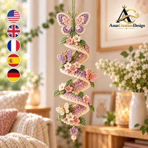 May include: A hanging crochet spiral ornament featuring a lavender and cream ribbon design adorned with small green leaves, pink and white flowers, and several butterflies. The piece includes gold-toned accents and a single white pearl at the bottom.