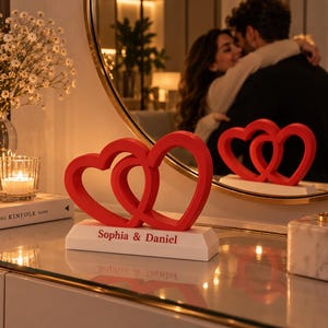 May include: A red decorative sculpture featuring two interlocking heart shapes mounted on a white rectangular base. The base is printed with the text Sophia & Daniel in a red font. The object is positioned on a reflective surface.