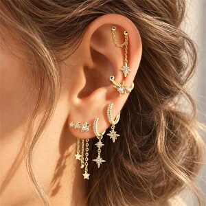 May include: A set of gold-toned earrings featuring celestial motifs including stars and a crescent moon. The collection includes a chain-linked cartilage earring, a star-shaped ear cuff, and multiple hoop earrings adorned with small clear stones.