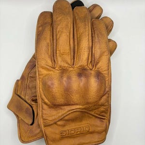 May include: A pair of tan leather gloves featuring a reinforced knuckle guard and a debossed SIDRIO logo on the wrist area. The gloves have a textured, worn-in appearance with visible stitching along the fingers and a secure closure strap at the base.