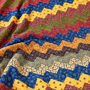 May include: A quilted textile featuring a repeating chevron pattern composed of various fabric strips. The design incorporates a vibrant colour palette of yellow, red, blue, green, and cream, with each section displaying distinct floral and geometric prints.