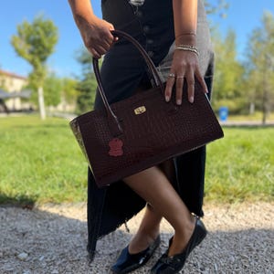 Peut inclure: Un sac à main bordeaux foncé avec une texture gaufrée façon crocodile, doté d'une fermeture à tourniquet en métal doré et de deux anses. Une petite étiquette rectangulaire en cuir rouge est fixée à l'avant du sac de forme rectangulaire.