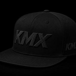 May include: A solid black baseball cap featuring the letters KMX embroidered in a matching black thread on the front panel. The same KMX lettering is also visible on the side of the cap. The design includes a flat brim and a structured crown.