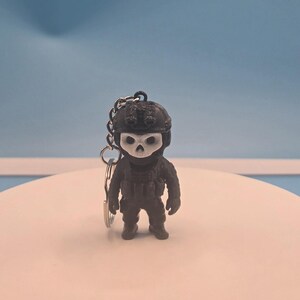 May include: A black keychain featuring a small figurine of a soldier wearing a tactical helmet and a white skull mask. The figure is equipped with a vest and gear, and is attached to a silver-colored metal split ring for keys and accessories.